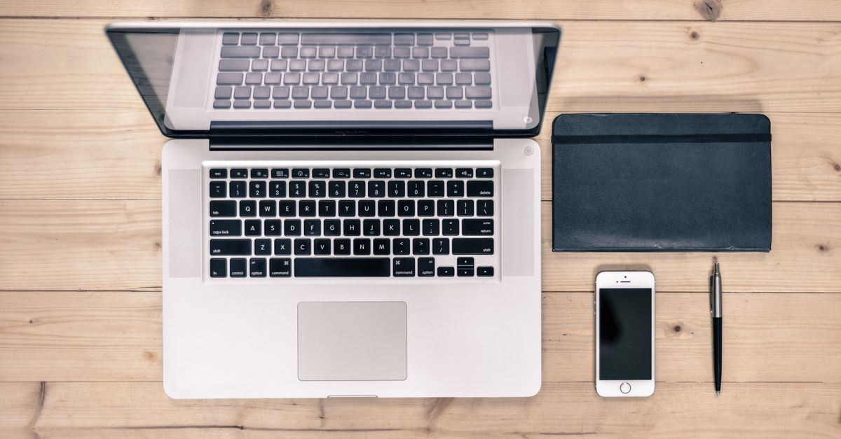 A laptop, notebook, phone and pen arranged neatly on a wooden desk, seen from above