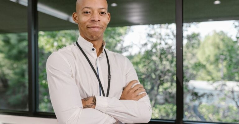 A man in a white shirt and lanyard standing with his arms folded by an office window