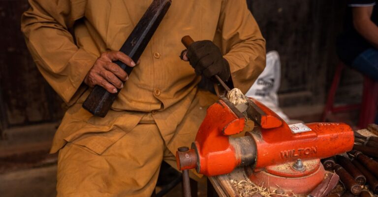 A worker in a yellow jacket using a chisel and a bench vice in a workshop