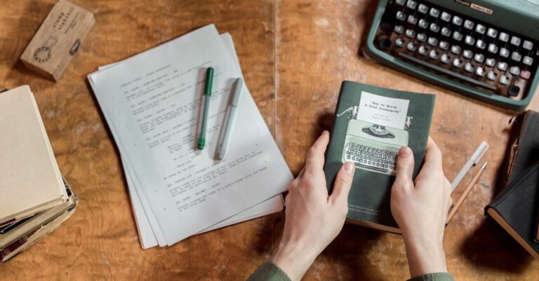 Hands holding a notebook on a wooden desk, beside a typewriter and loose manuscript pages