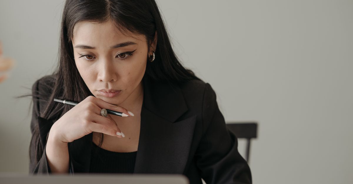 A woman holding a pen and thinking while working at a laptop