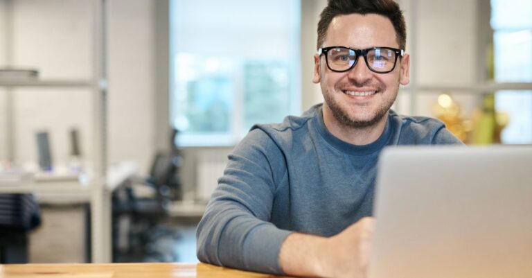 A smiling man in glasses working at a laptop in an open-plan office
