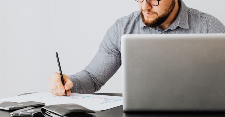 A man writing in a notebook beside a laptop