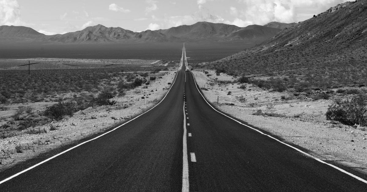 A straight empty road running through desert toward distant mountains, in black and white