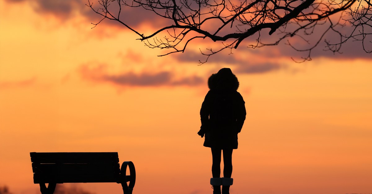 A serene silhouette of a person standing next to a bench against a vibrant sunrise sky.