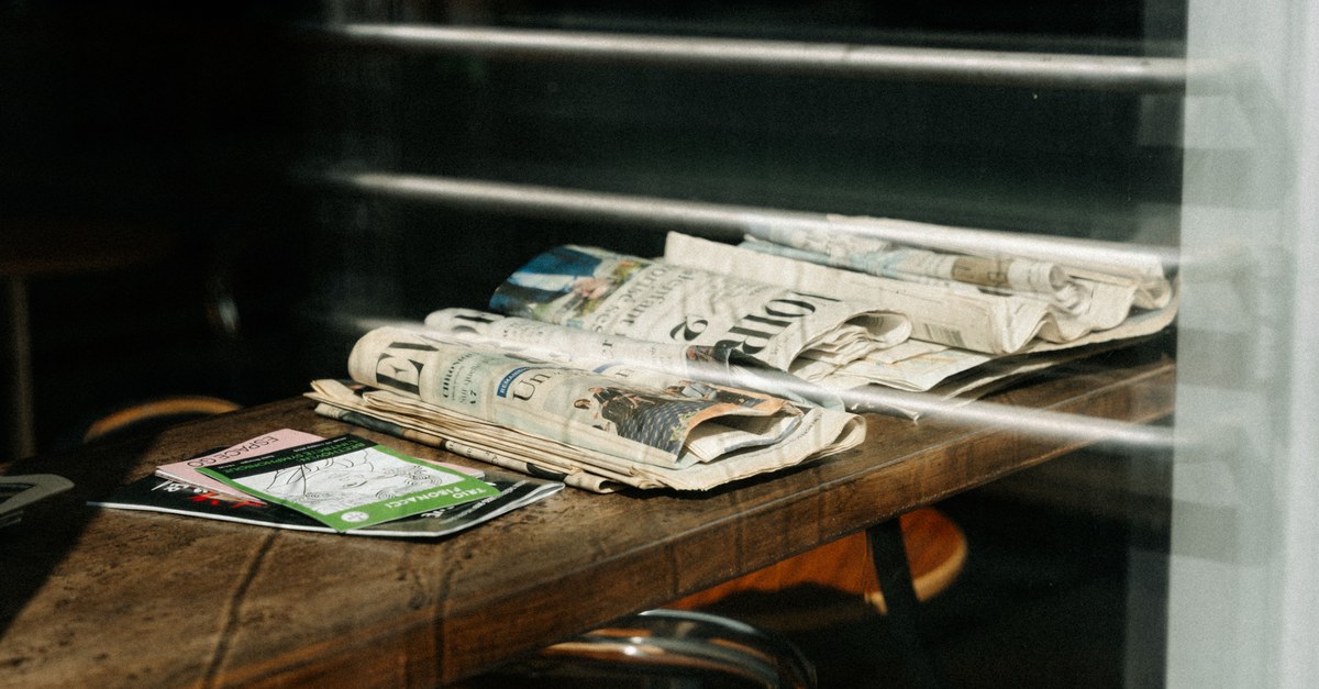 Folded newspapers and magazines stacked on a wooden cafe table beside a sunlit window