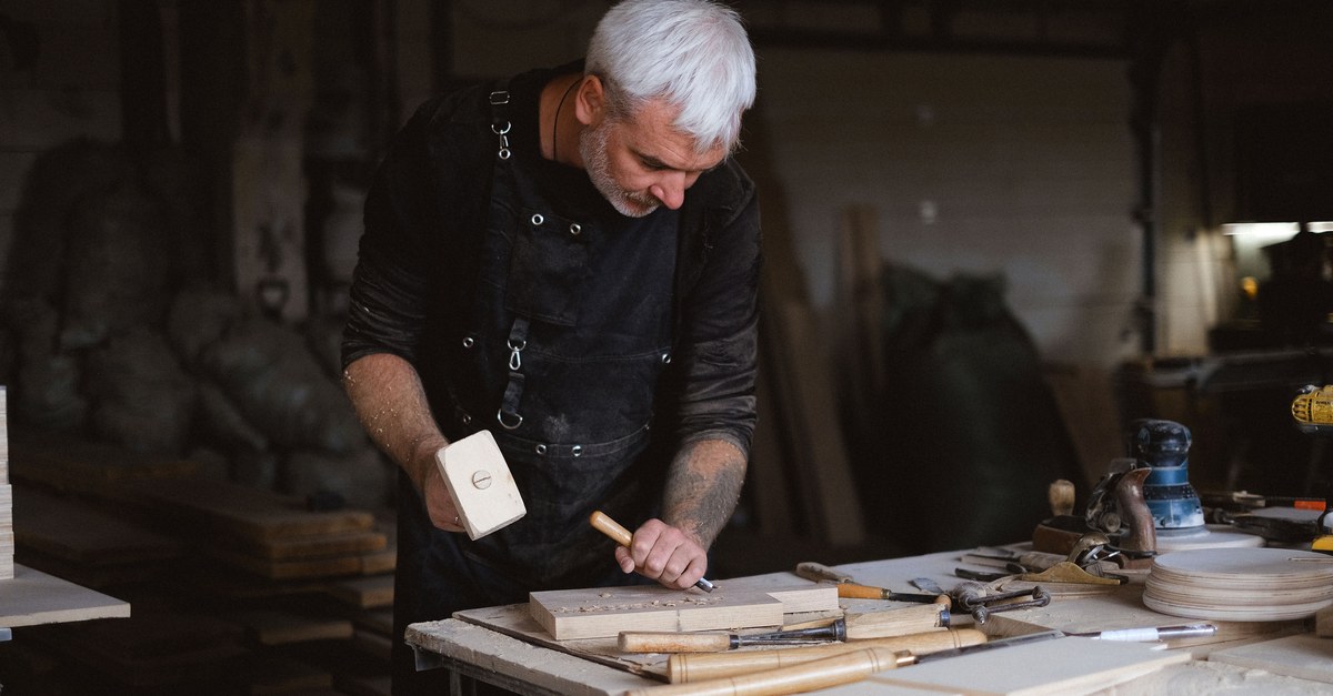 A gray-haired woodworker in a black apron bends over a workbench, mallet raised in one hand and chisel in the other, with hand tools laid out in front of him