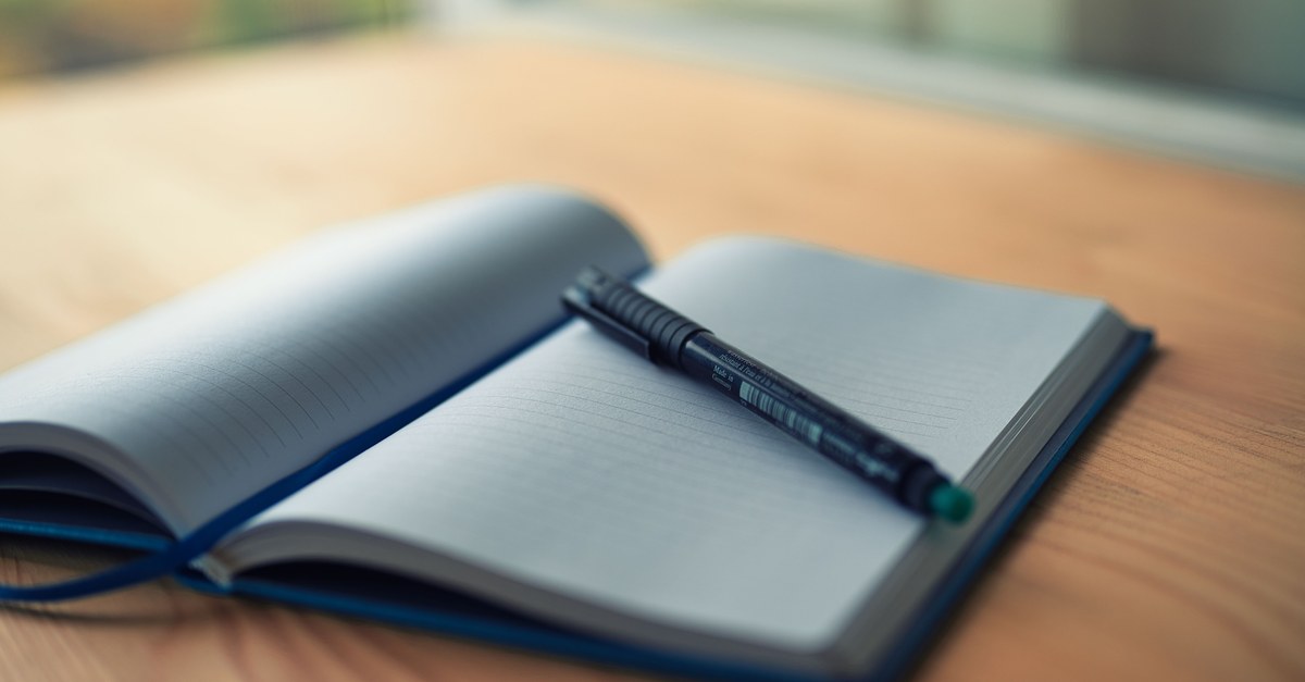 An open blank notebook with a pen resting on it, on a wooden desk in soft window light