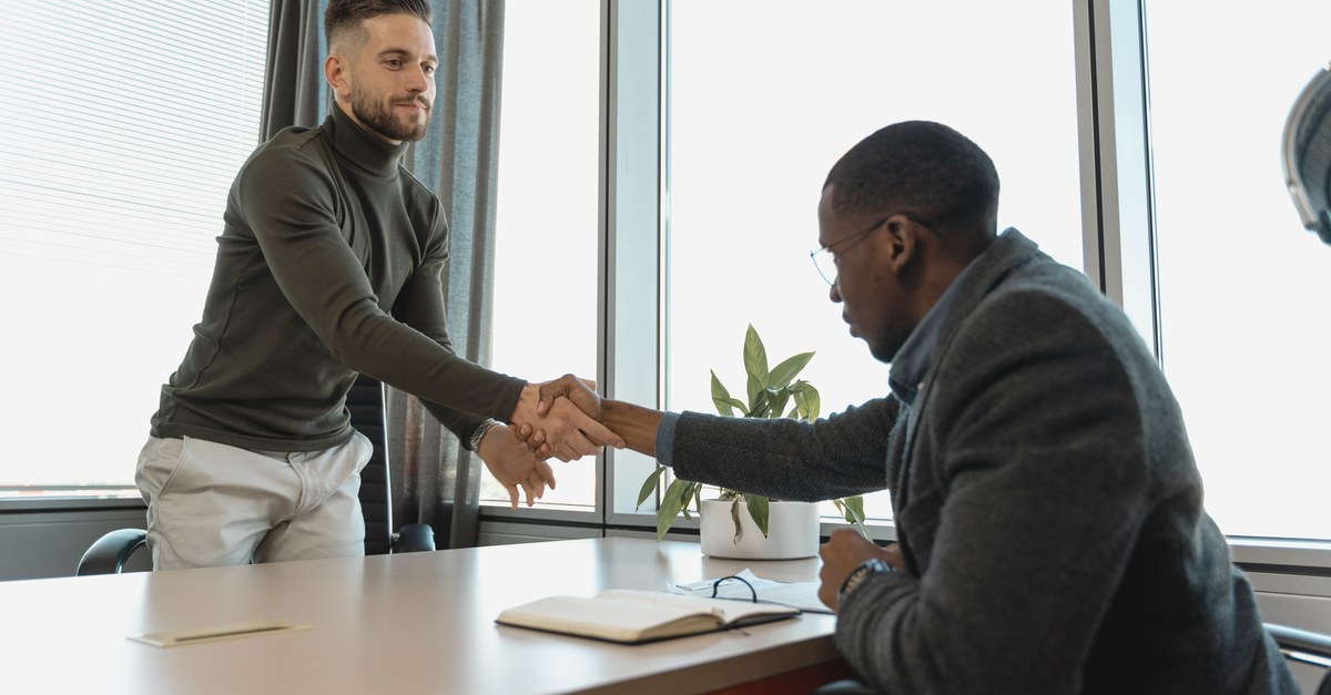 A man in a dark green sweater and a man in a gray jacket shaking hands across a pale desk by tall bright windows