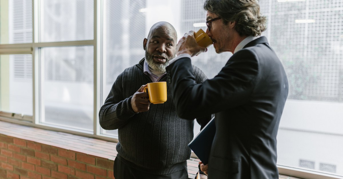 Two men in an office hallway talking over coffee by a tall window, one holding a yellow mug and a folder