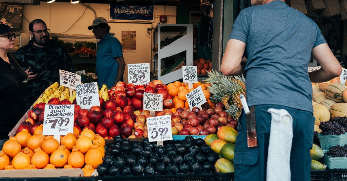 Handwritten price signs on stacks of plums, apples, oranges and pears at a busy outdoor produce stall